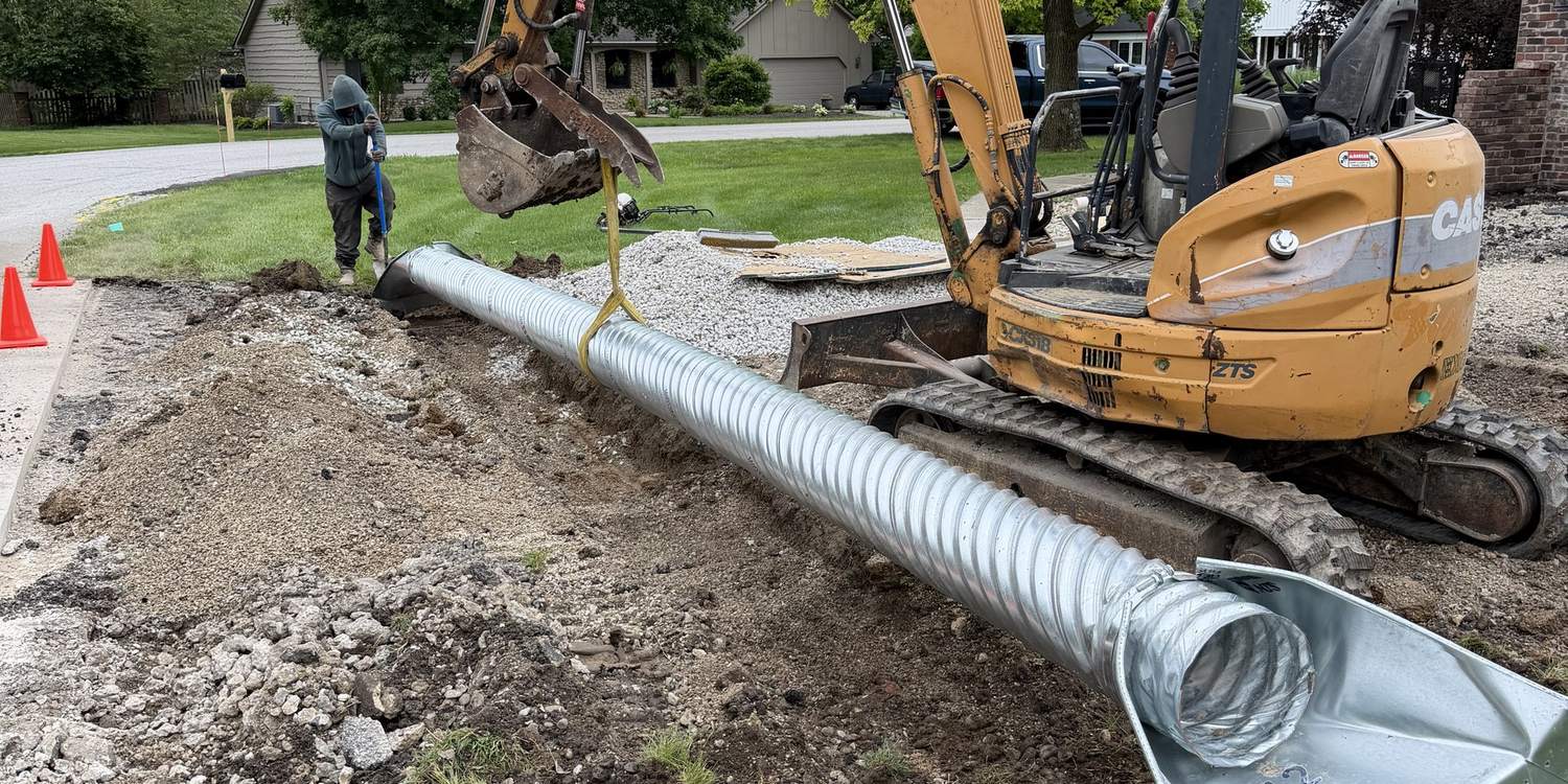 Corrugated metal culvert staged beside an open trench with a compact excavator