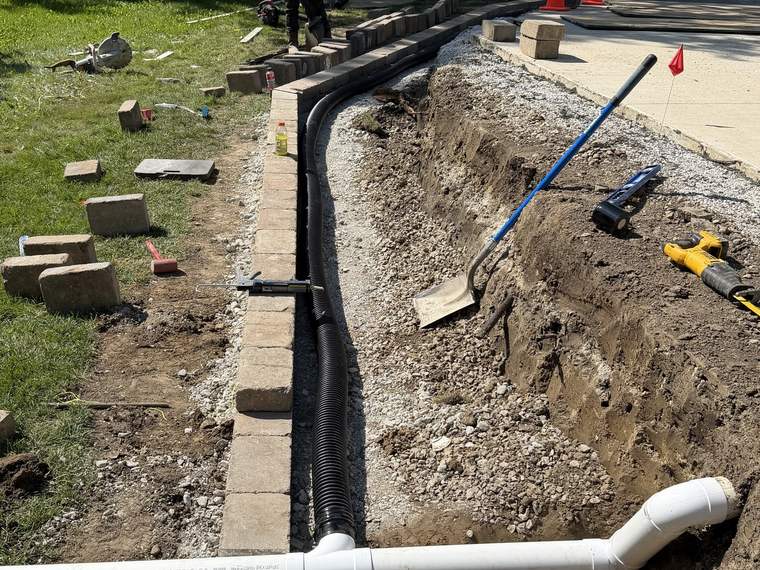 Downspout collection piping installed in an open trench beside a paved walk
